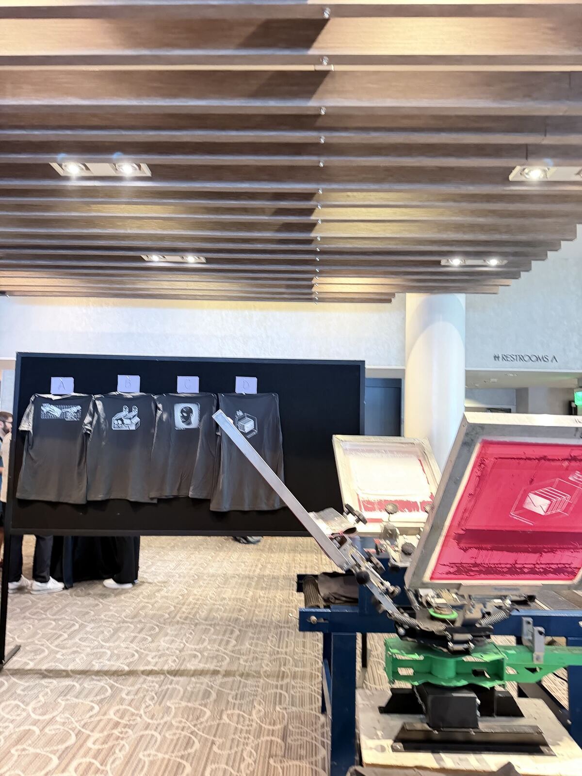 Display board of four sample designs hanging beside a live press at a carpeted hotel venue